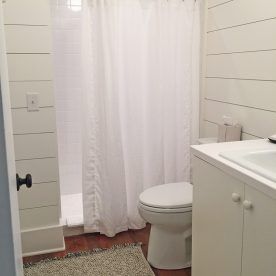 Small bathroom with white shiplap walls, shower, toilet, and vanity; wood floor; light rug.