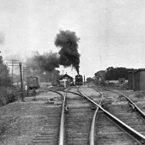 Black and white photo of a steam train at a railway station with smoke billowing into the sky.