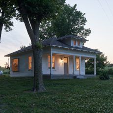 White house with a porch, lit windows, tree in foreground, grassy yard at dusk.