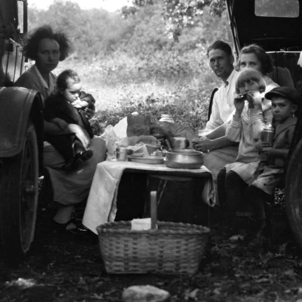 Family picnic, circa 1930s, around a car trunk table. People eating, seated, in a wooded area; basket in foreground.