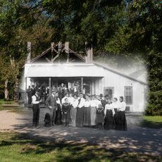 Group of people in front of a white building, possibly a school. Trees surround them.