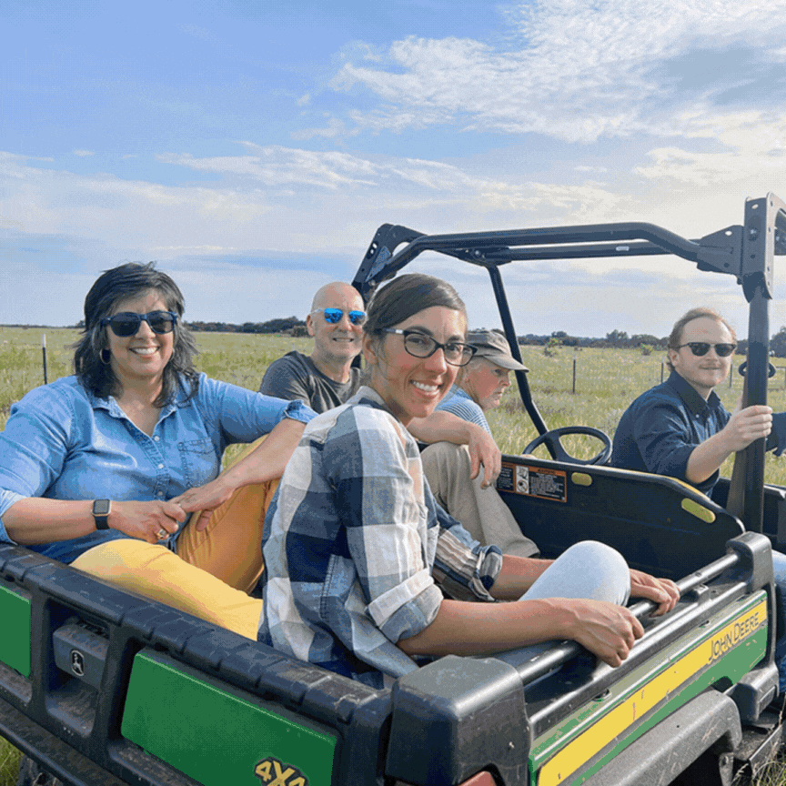Five people ride in a green utility vehicle across a grassy field under a blue sky.