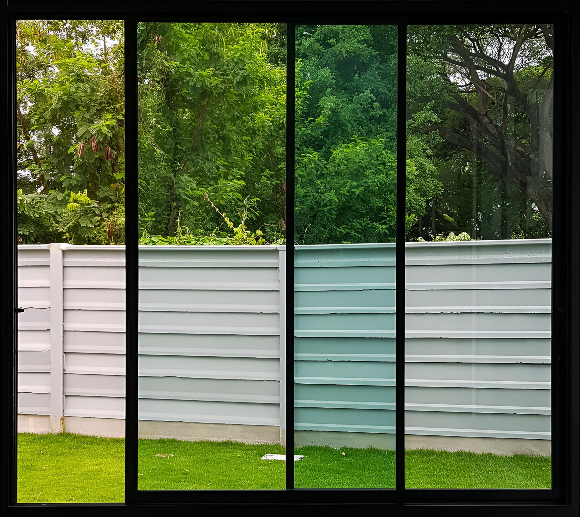 Four-panel window framing a backyard view: green trees, white fence, and lawn.