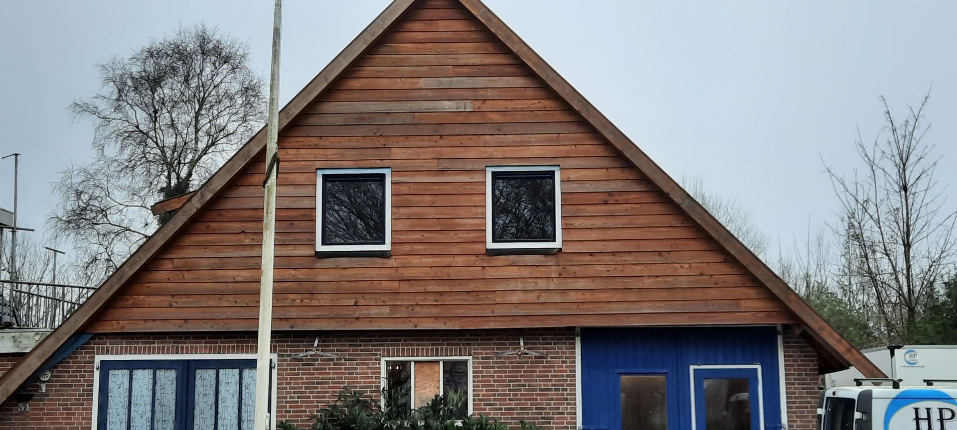 Houten huis met steil dak, twee kleine ramen op de bovenverdieping en blauwe gevelbekleding onder een bewolkte hemel.