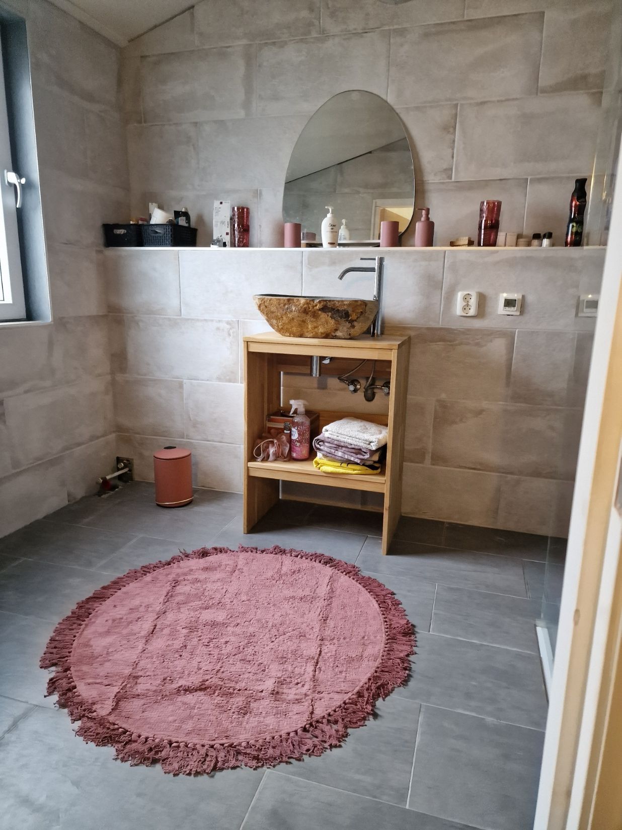 Kleine badkamer met stenen muren, een plankje bij de wastafel en een roze rond vloerkleed op een grijze vloer.