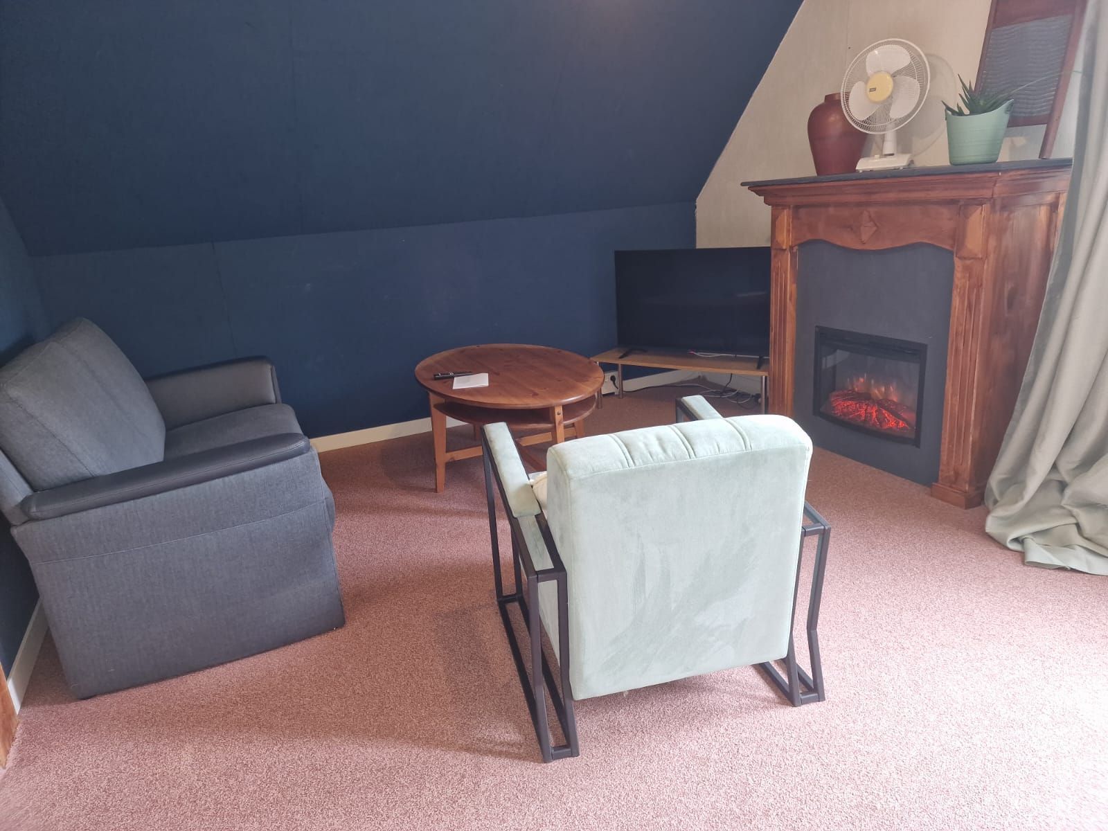 Gezellige zolderwoonkamer met grijze bank, twee fauteuils, salontafel en open haard.