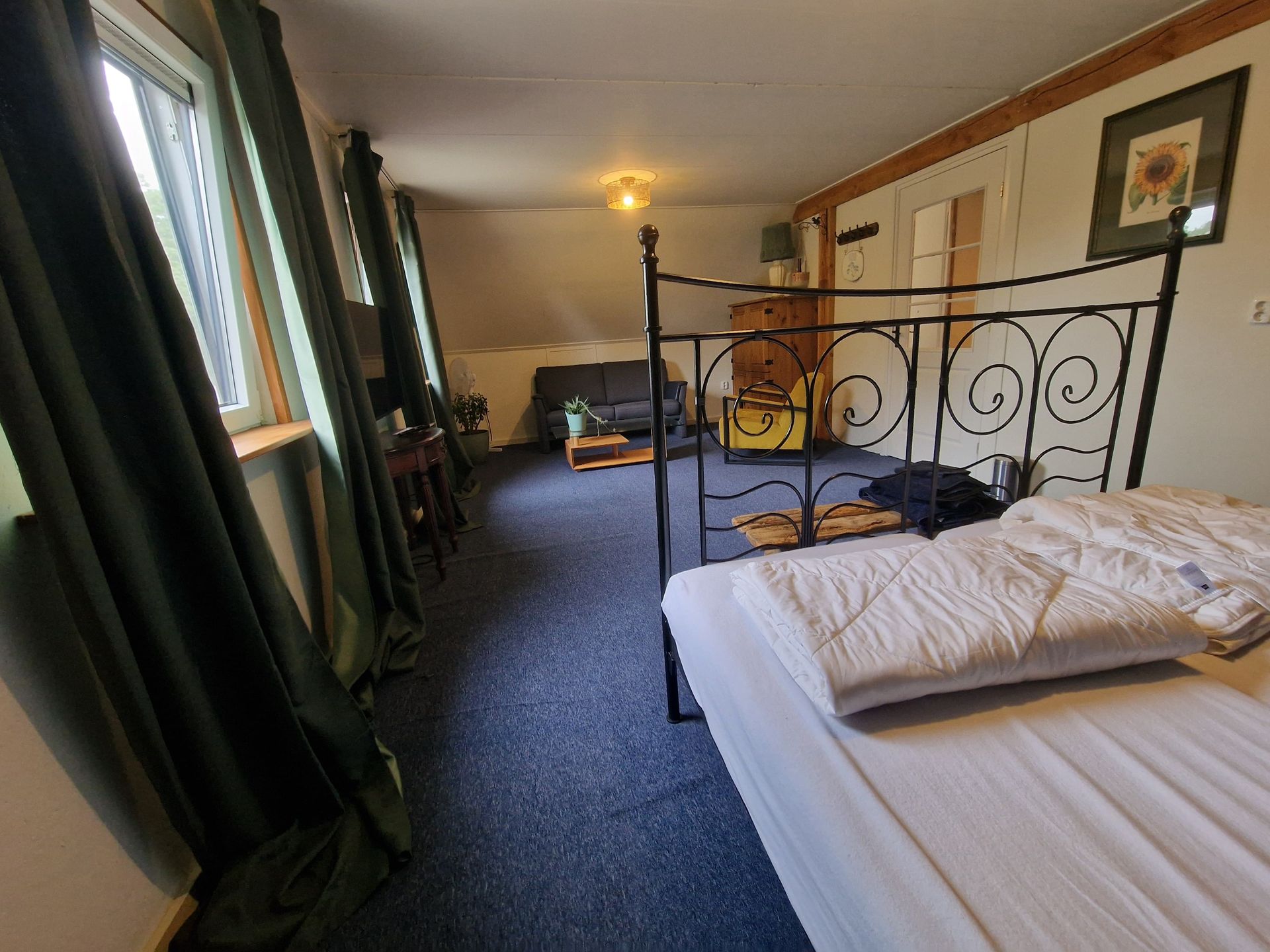 Lange slaapkamer met een zwart smeedijzeren bed, wit beddengoed, groene gordijnen en een gele stoel bij het raam.