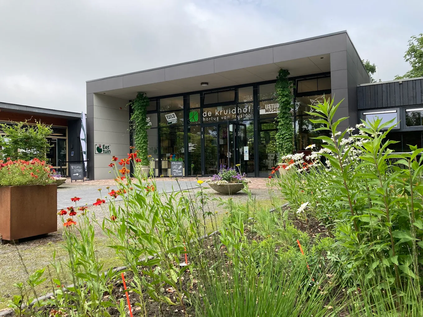 Moderne gebouwingang met glazen deuren, weelderige tuinplanten en bloemperken aan de voorzijde.