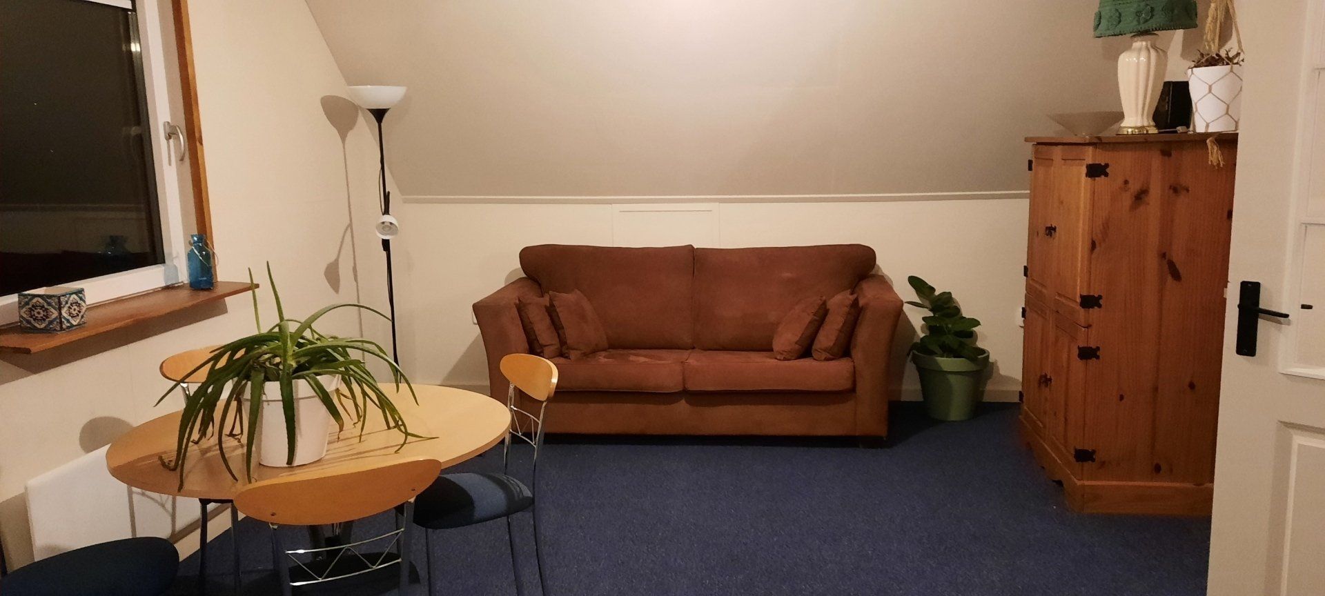 Gezellige woonkamer met bruine bank, houten kast, ronde tafel, blauw tapijt en een staande lamp.