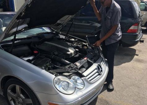 Mechanic inspecting a silver Mercedes engine with the hood open in a sunny outdoor setting.