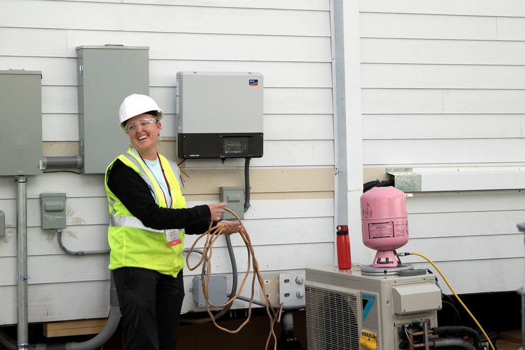 A woman in a hard hat and safety vest is working on an air conditioner.