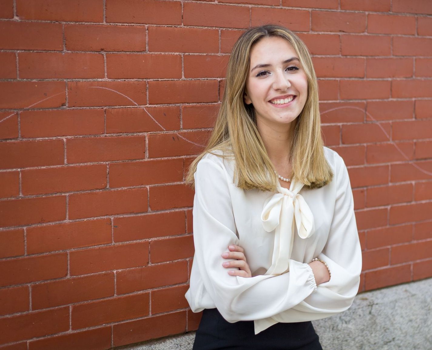 A woman is standing in front of a brick wall with her arms crossed and smiling.