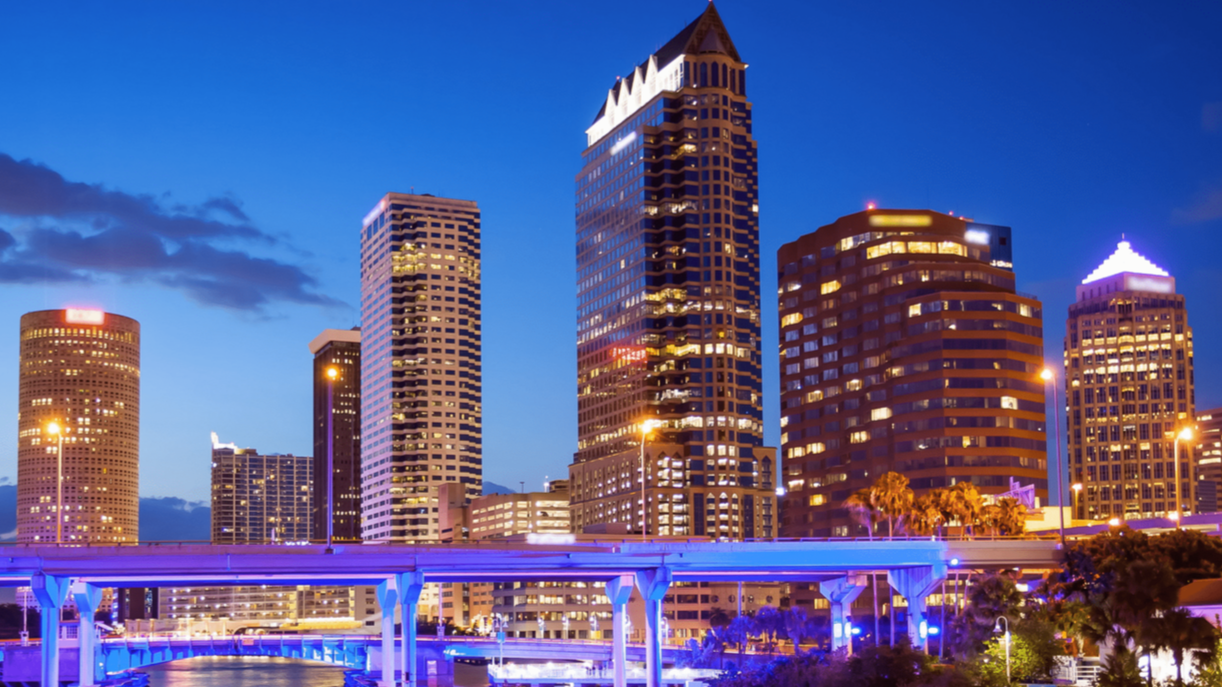 Nighttime view of Tampa, Florida skyline with lit skyscrapers and a blue bridge.