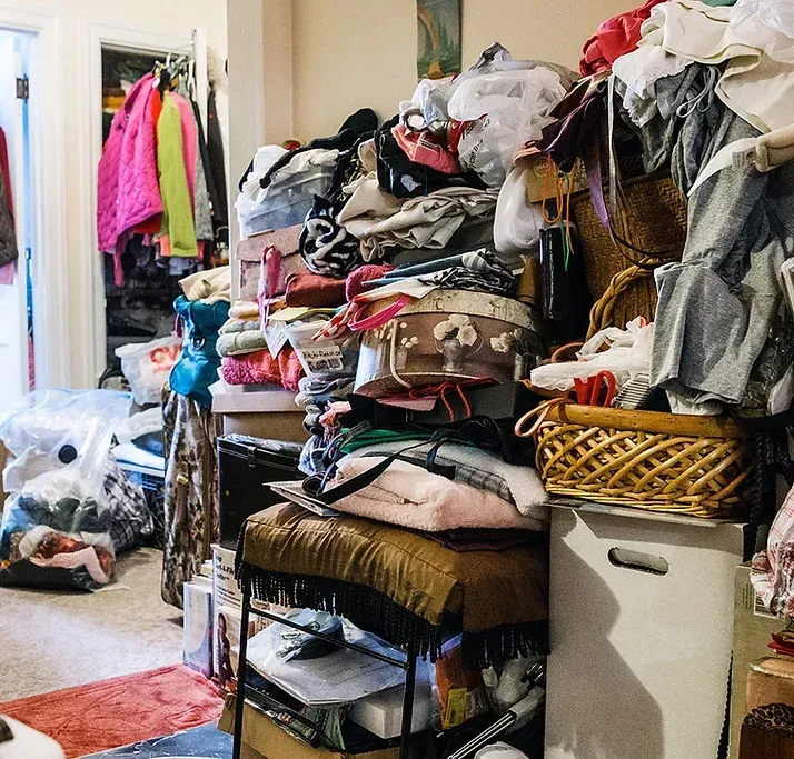 A cluttered room with piles of clothes, boxes, and baskets. Clothes hang in a partially visible closet.