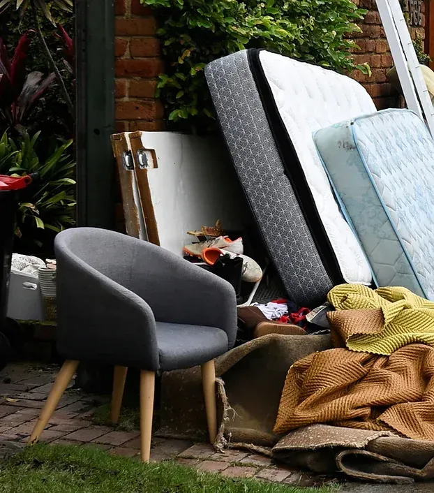 A gray armchair sits in front of a pile of discarded furniture: mattresses, a door, and blankets, near a brick wall.