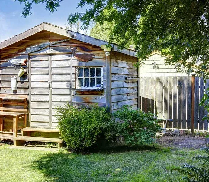 Weathered wooden shed with small window and door, surrounded by greenery and a grassy yard.