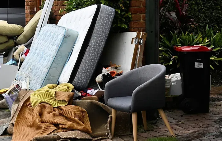 Pile of discarded items, including mattresses, chair, rug, and trash bin, on a curb.