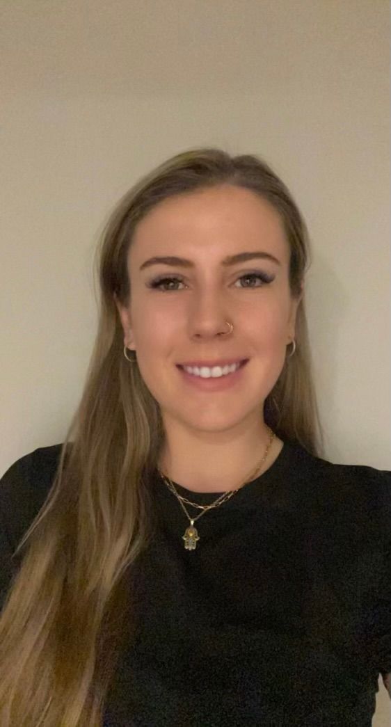 Smiling person with long blonde hair wearing a black top against a plain beige wall