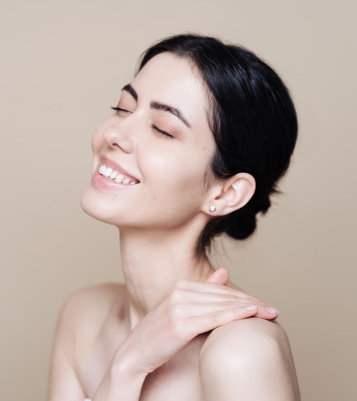 Smiling woman with closed eyes gently touching her shoulder against a beige background