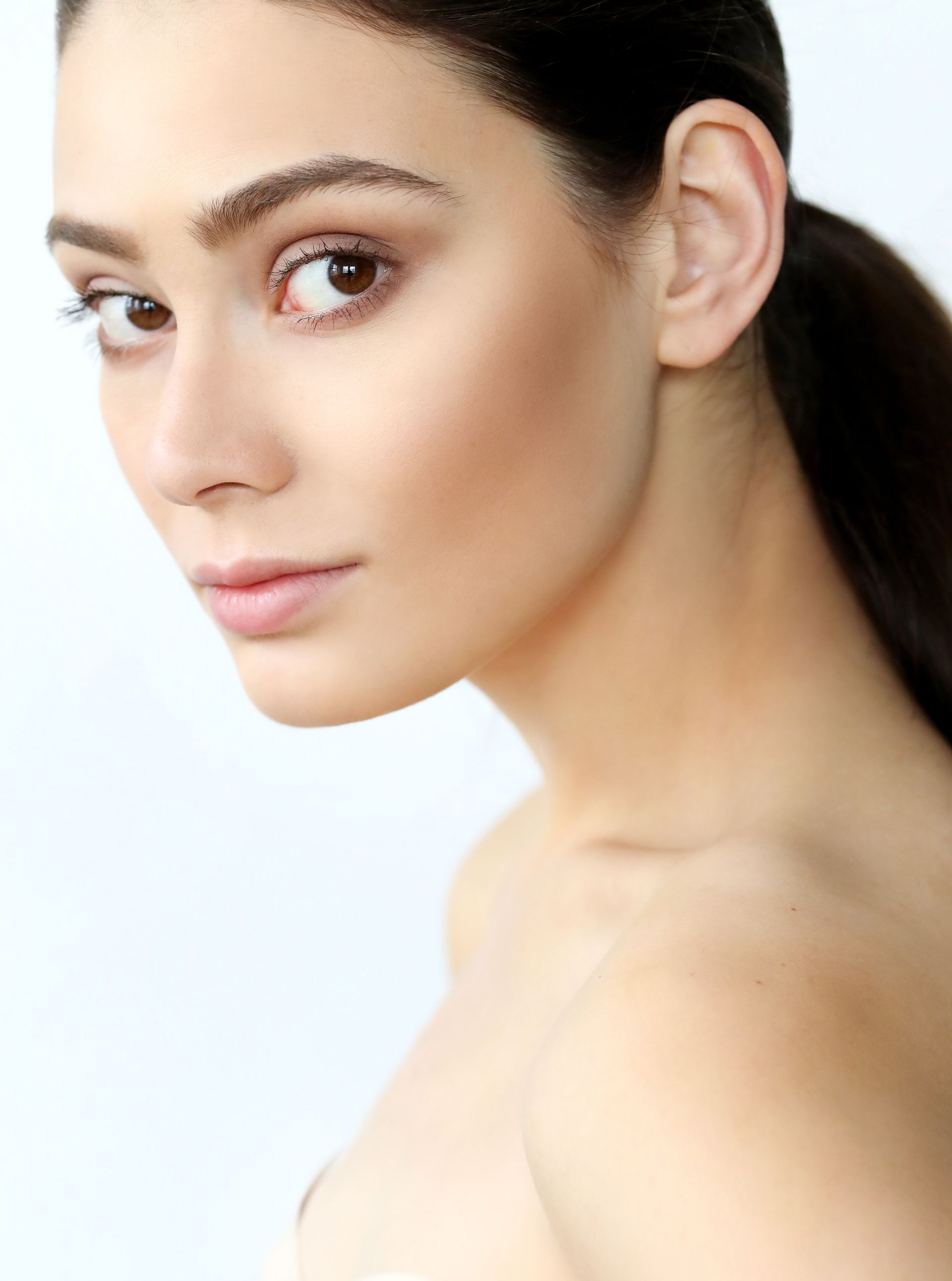 Close-up portrait of a woman with dark hair, looking over her shoulder against a light background