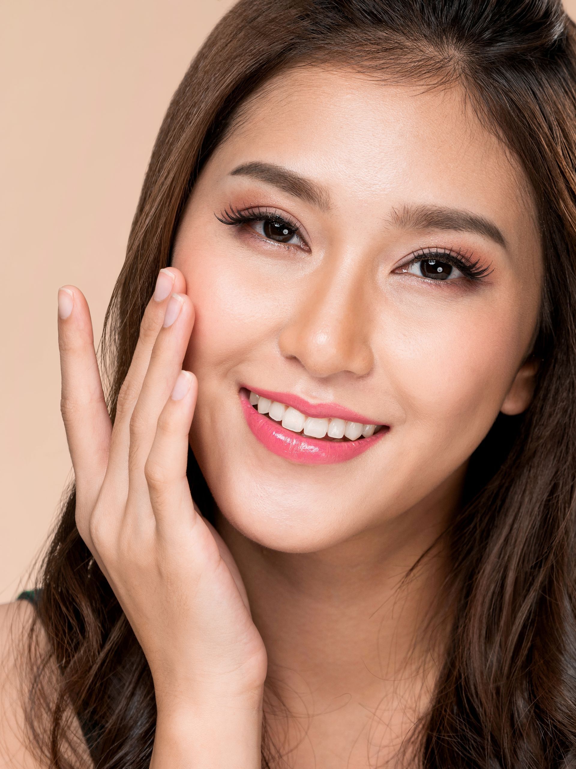 Smiling woman with long brown hair touching her cheek against a beige background