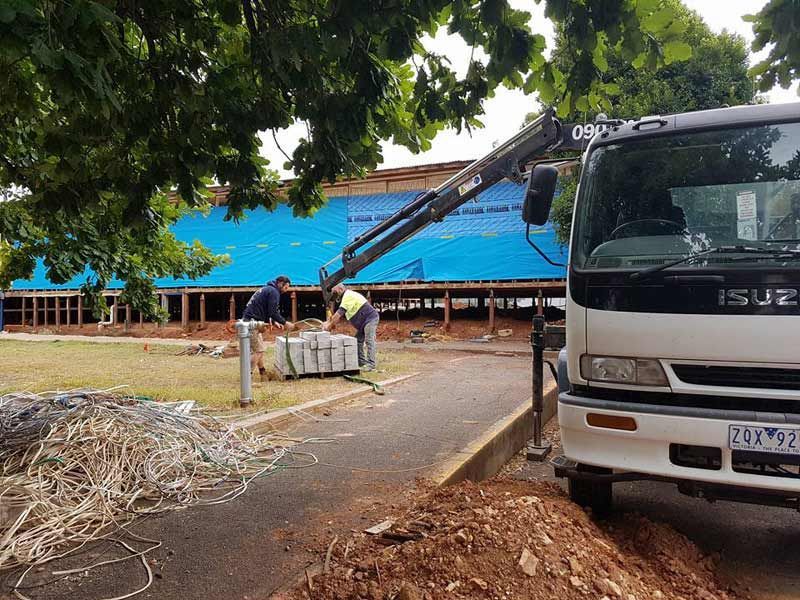 Worker using a crane to move debris beside a white truck and blue building under trees