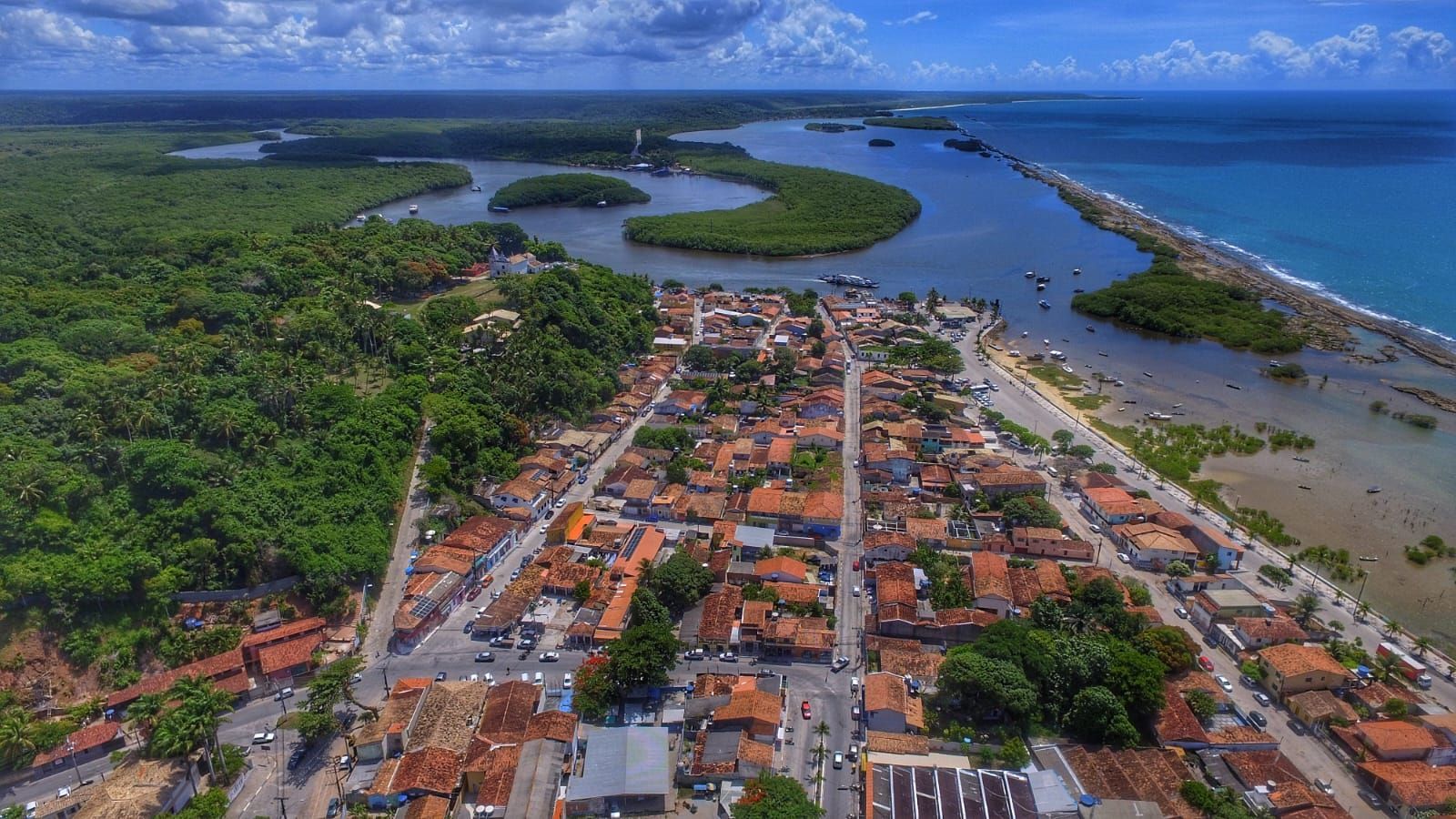 Vista aérea de uma cidade costeira junto a um rio sinuoso, com densa floresta verde e litoral oceânico.