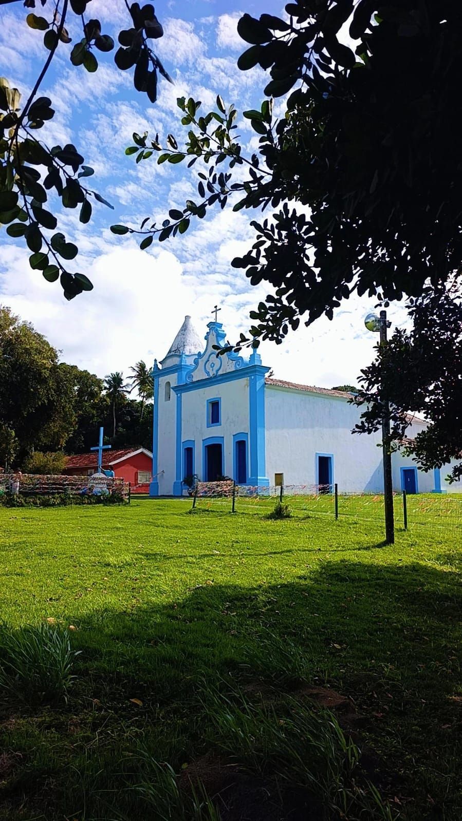 Igreja azul e branca em uma colina gramada sob um céu claro, emoldurada por galhos de árvores.