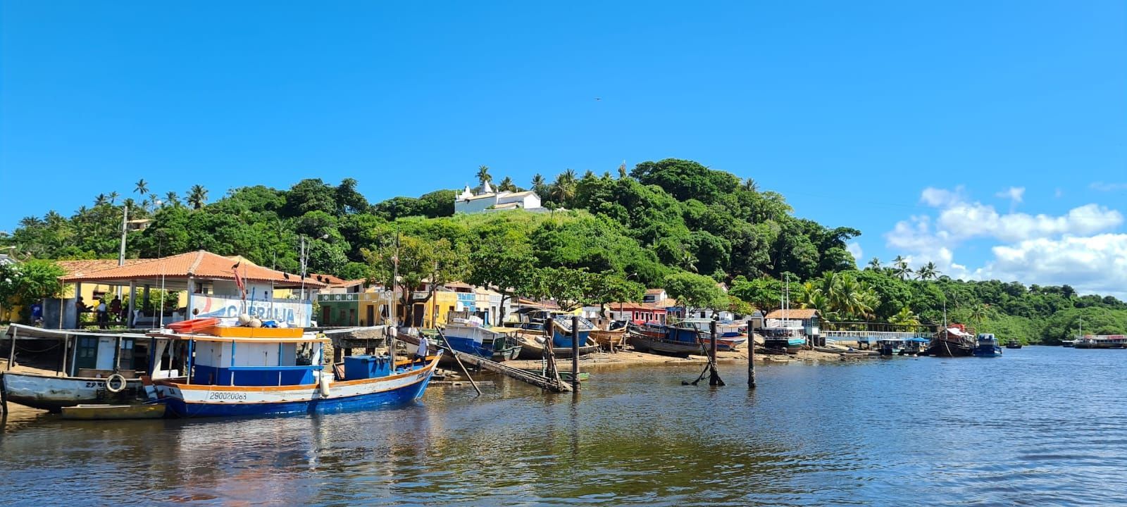 Barcos atracados ao longo de uma margem tranquila, ao lado de colinas verdejantes e céu azul.