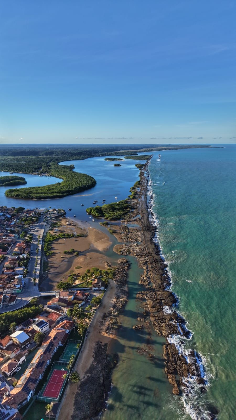 Vista aérea de uma cidade costeira junto ao oceano azul, com uma estreita faixa litorânea e um rio sinuoso sob um céu claro.