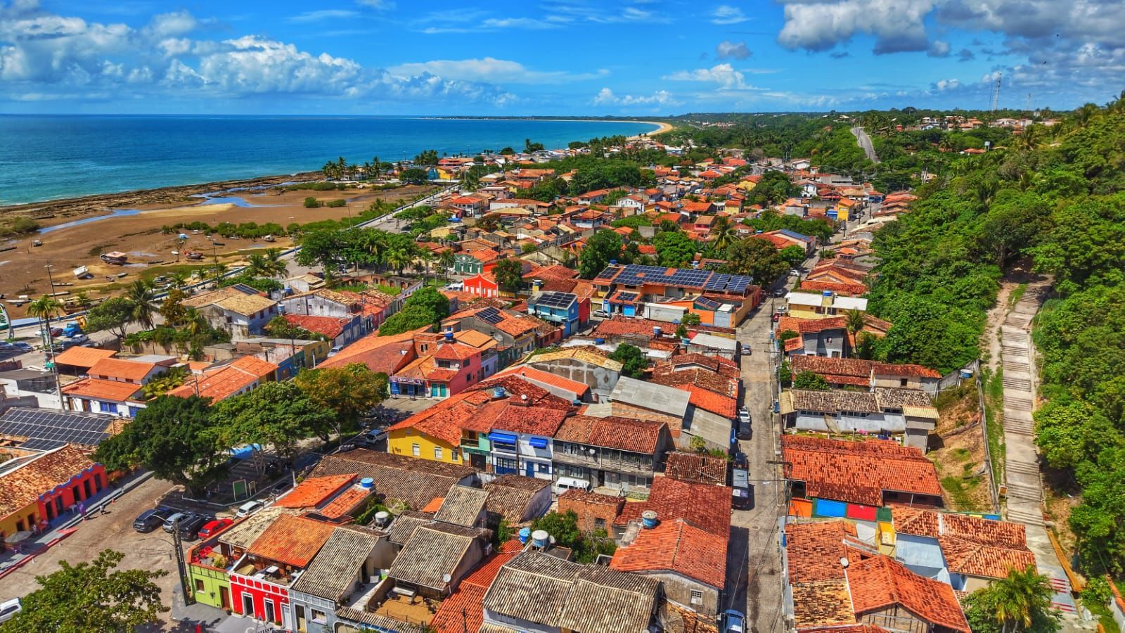Vista aérea de uma cidade costeira colorida com edifícios de telhados vermelhos junto ao mar azul e à costa rochosa.