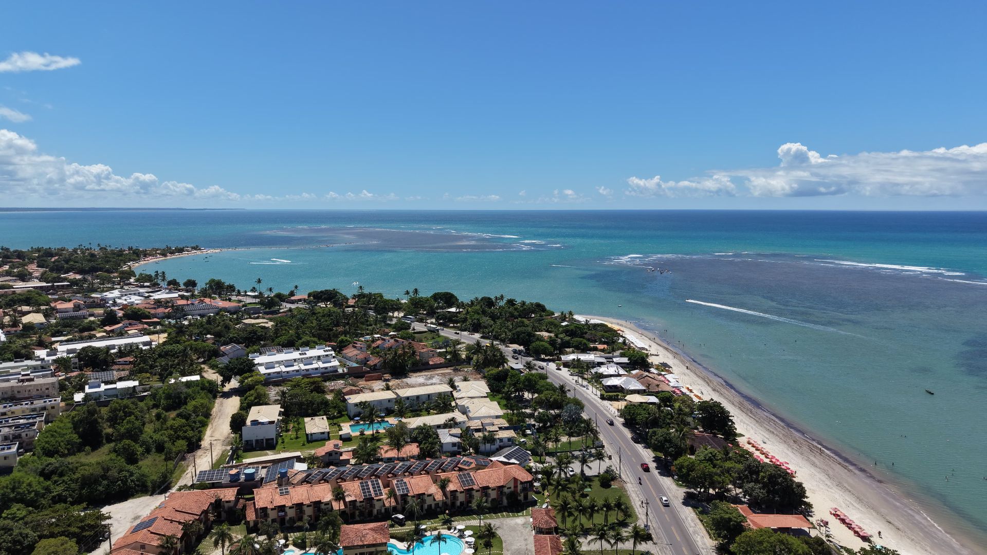 Vista aérea de uma cidade costeira com oceano turquesa, praia de areia e telhados sob um céu azul claro.