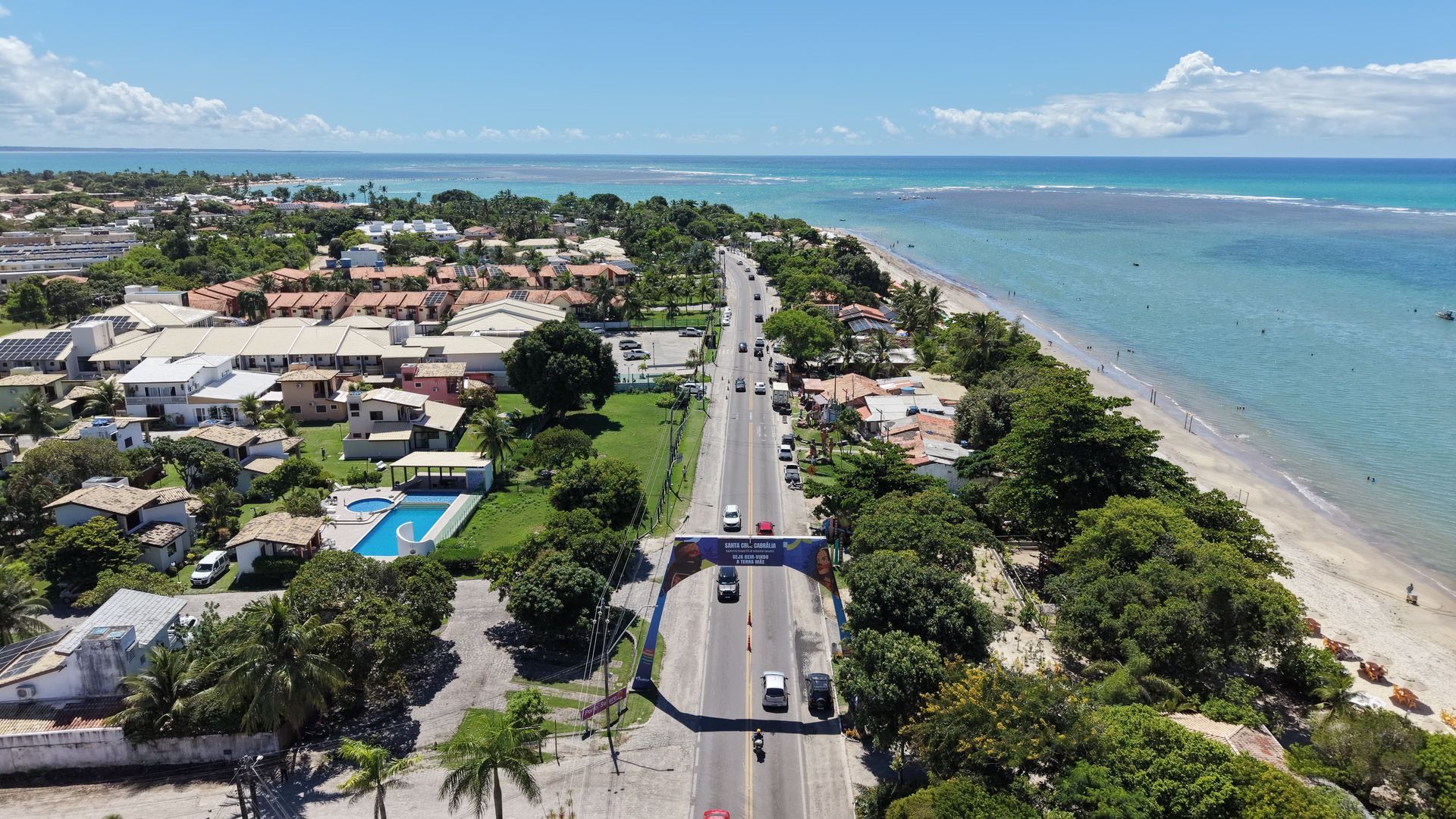 Vista aérea de uma estrada costeira junto ao oceano turquesa, ladeada por casas e árvores sob um céu limpo.