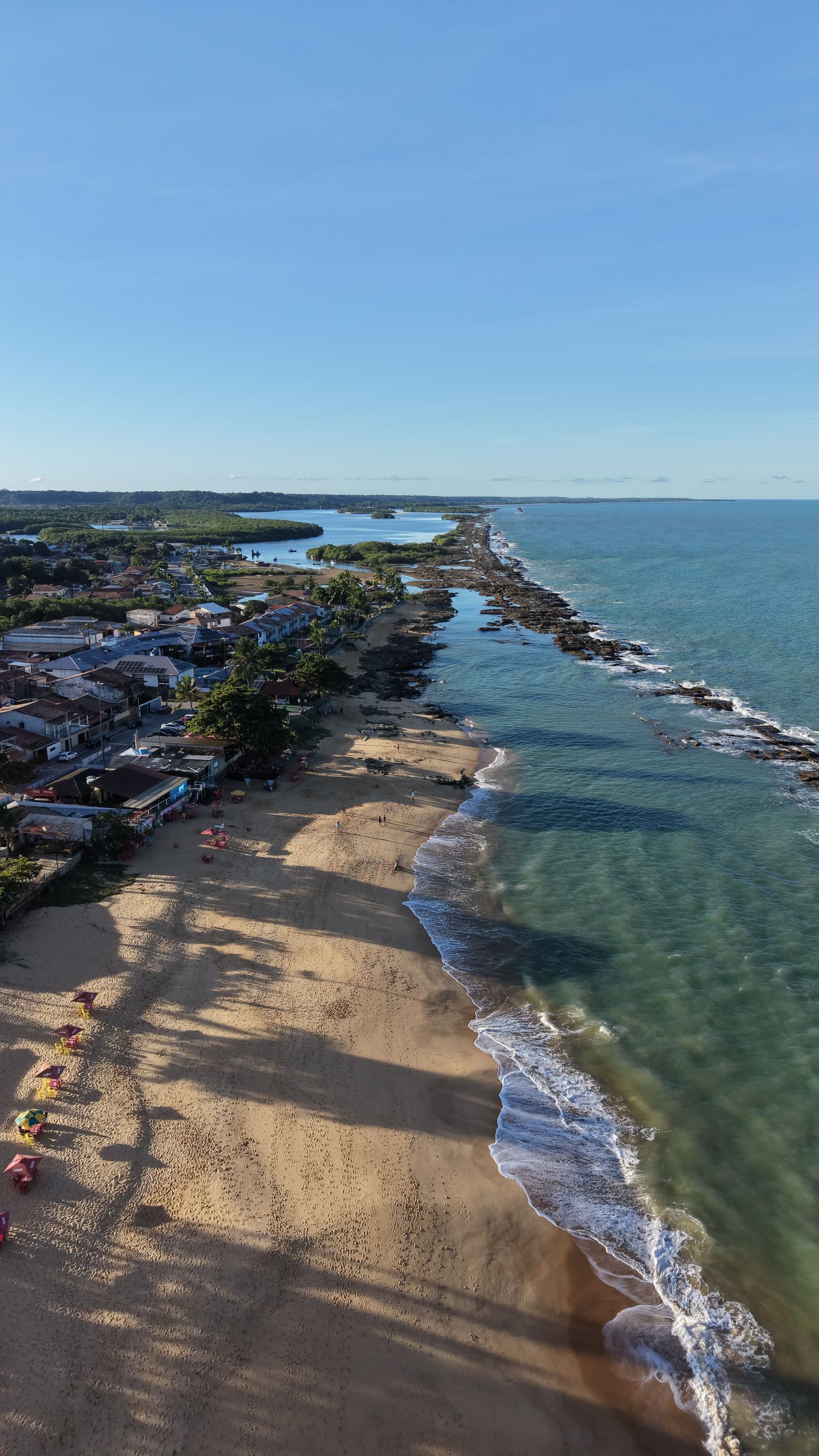 Vista aérea de uma praia de areia com água turquesa, quebra-mares rochosos e carros estacionados ao longo da costa.