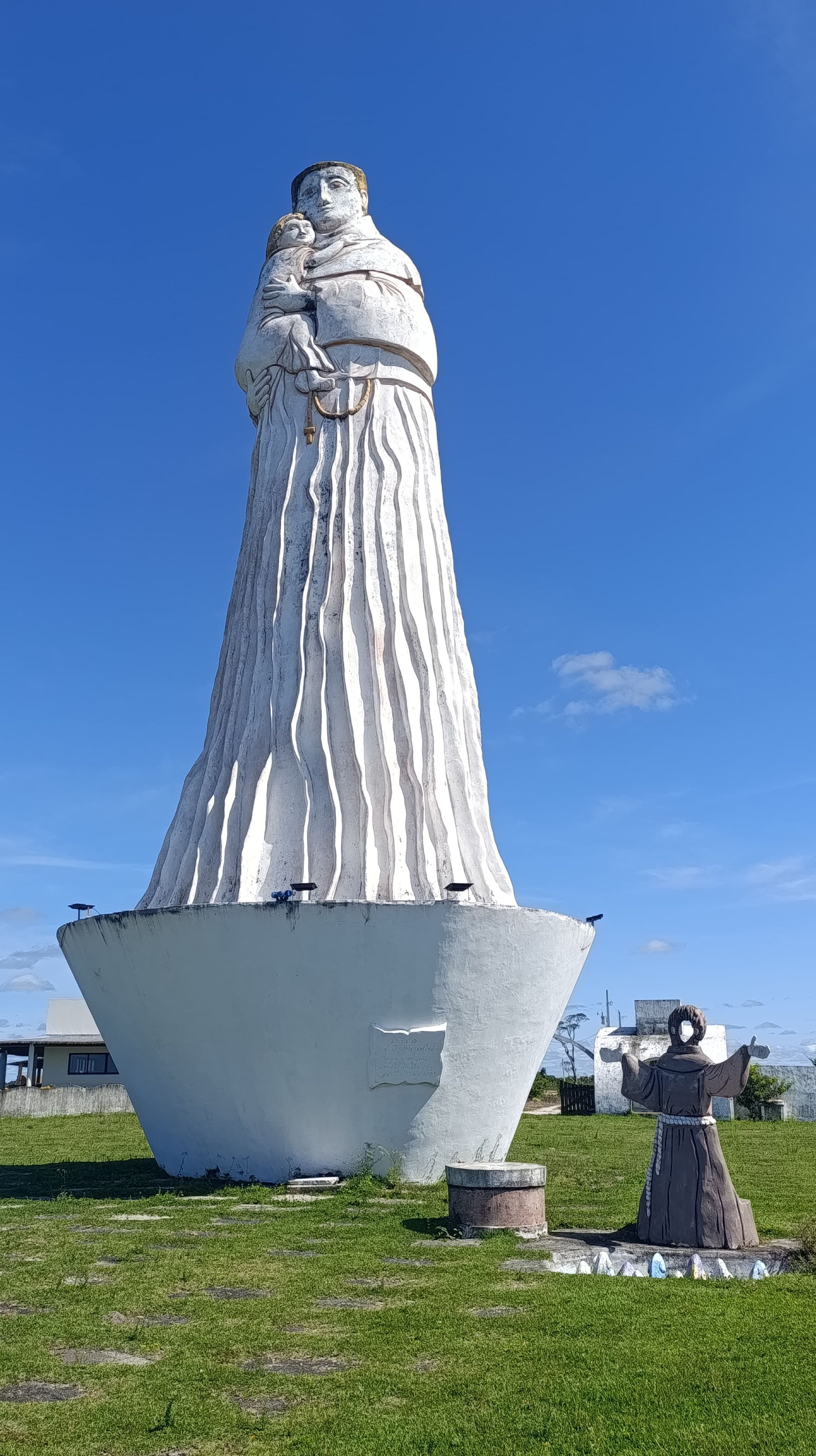 Estátua alta e branca sobre um pedestal em um campo gramado sob um céu azul claro.