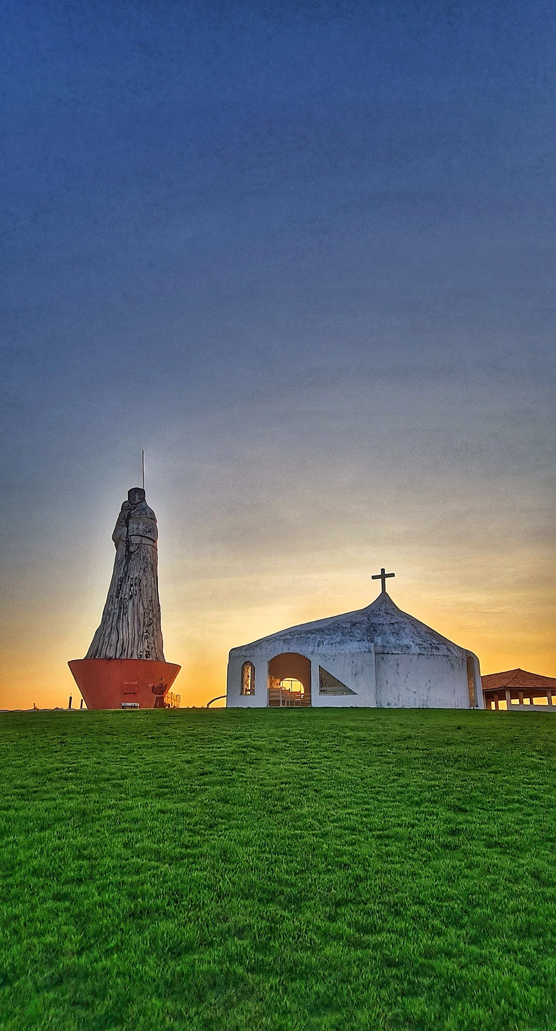 Campo gramado ao entardecer com uma capela branca iluminada e uma torre alta contra um céu azul-alaranjado.