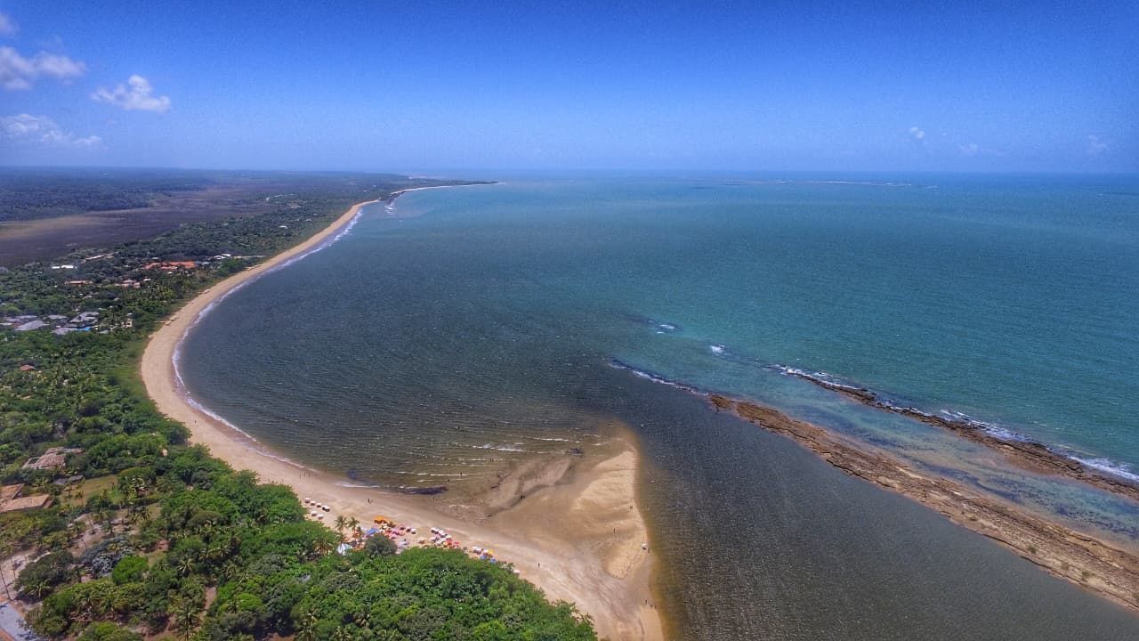 Vista aérea de uma praia de areia curva ao lado do oceano turquesa e da costa verde sob um céu limpo.