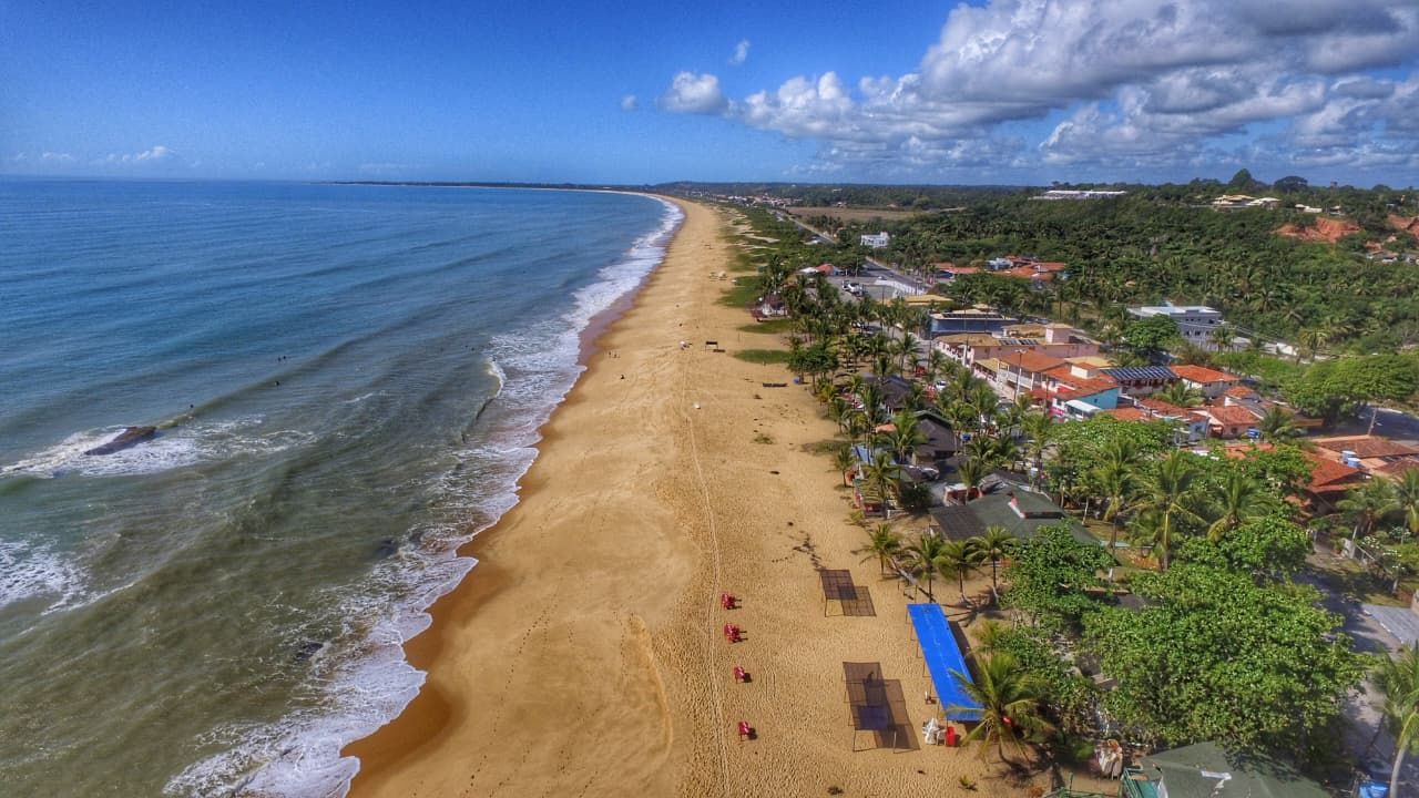 Vista aérea de uma extensa praia de areia junto ao oceano azul e uma cidade costeira sob um céu limpo.