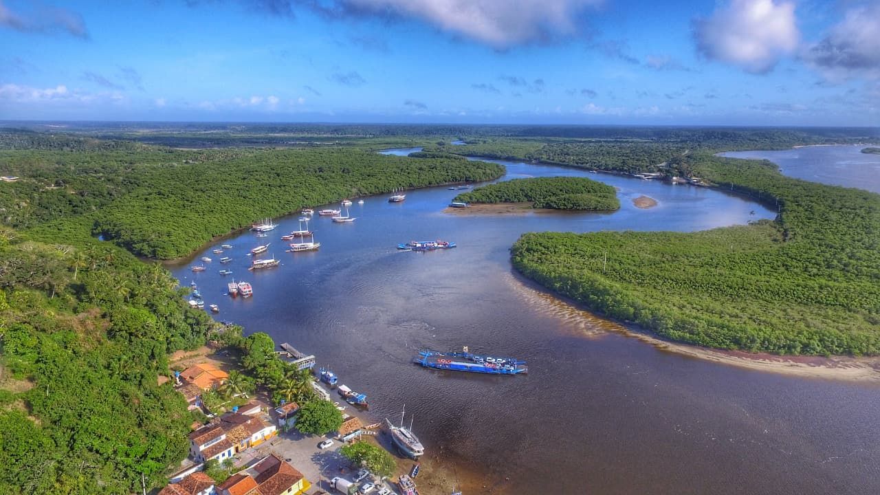 Vista aérea de um rio sinuoso atravessando uma densa floresta verde, com barcos e um pequeno povoado ribeirinho.