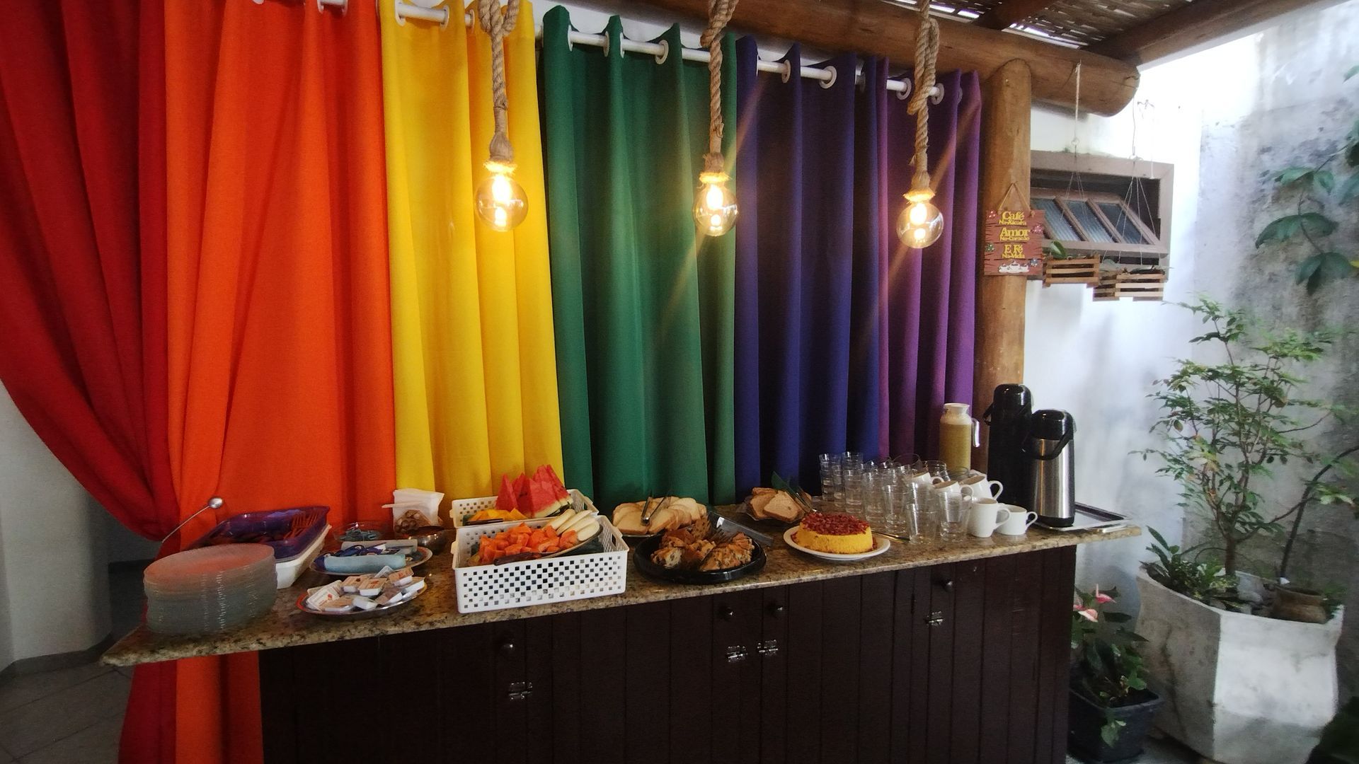 Painel de tecido colorido atrás de uma mesa de buffet com comidas e bebidas sob luzes pendentes.