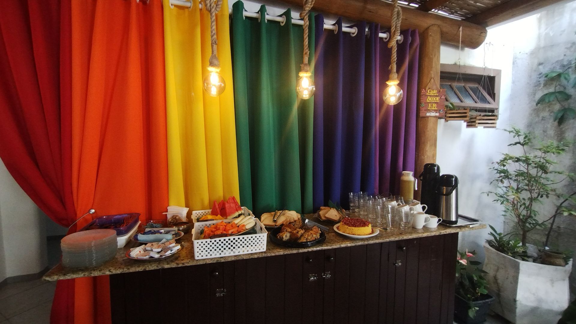 Um painel colorido e drapeado serve de pano de fundo para uma mesa de buffet com diversos petiscos e bebidas sob luzes pendentes.