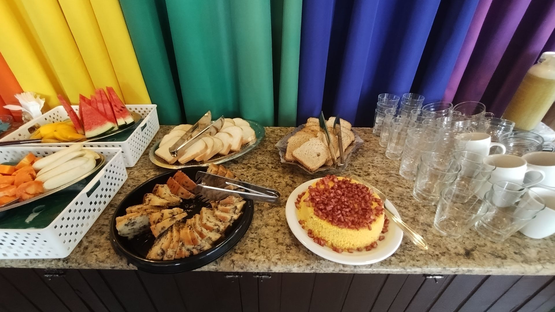 Mesa de buffet com sanduíches, doces, frutas, xícaras e um colorido tecido drapeado ao fundo.