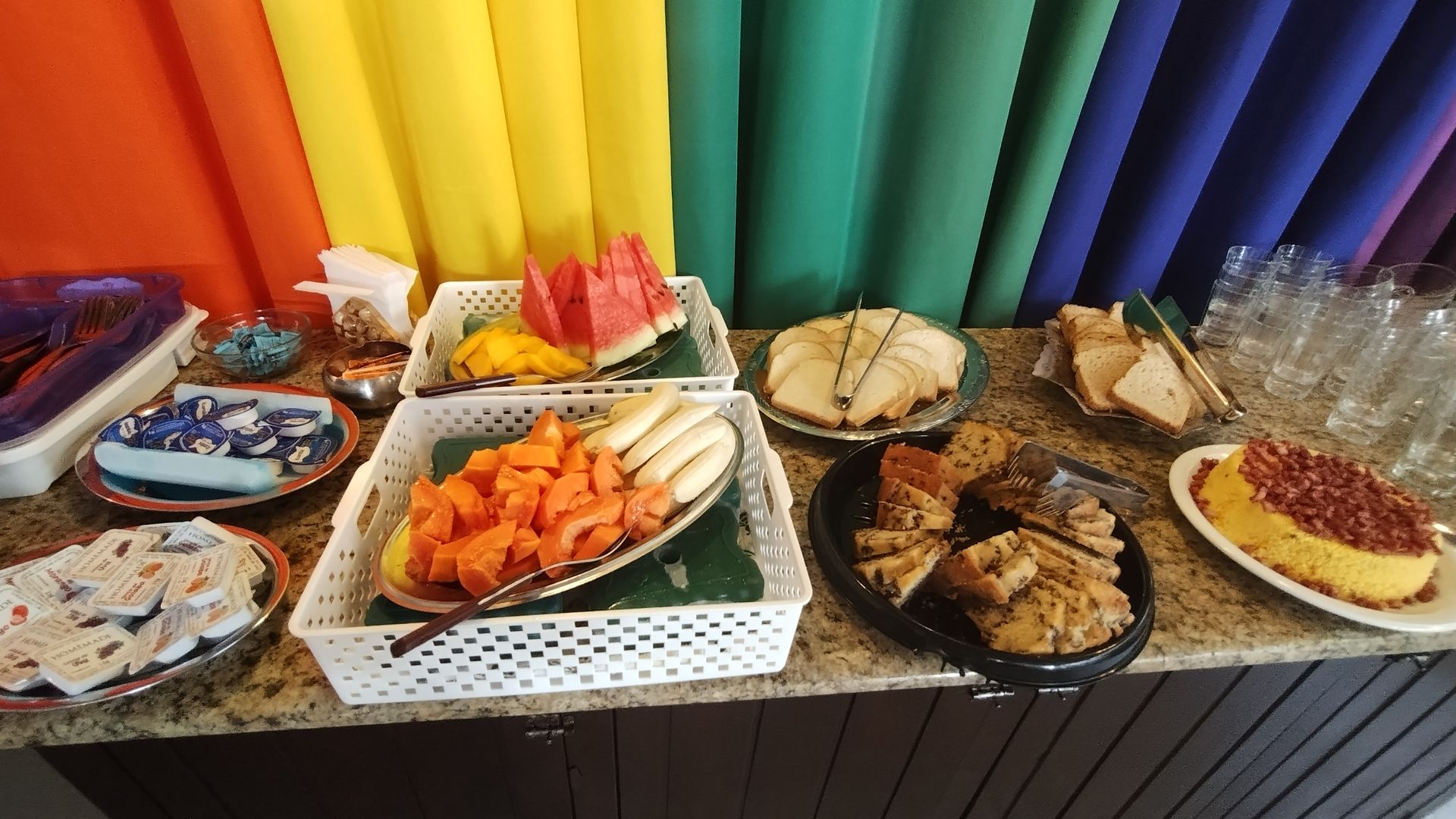 Mesa de bufê com uma variedade de doces e frutas em frente a cortinas coloridas em formato de arco-íris.