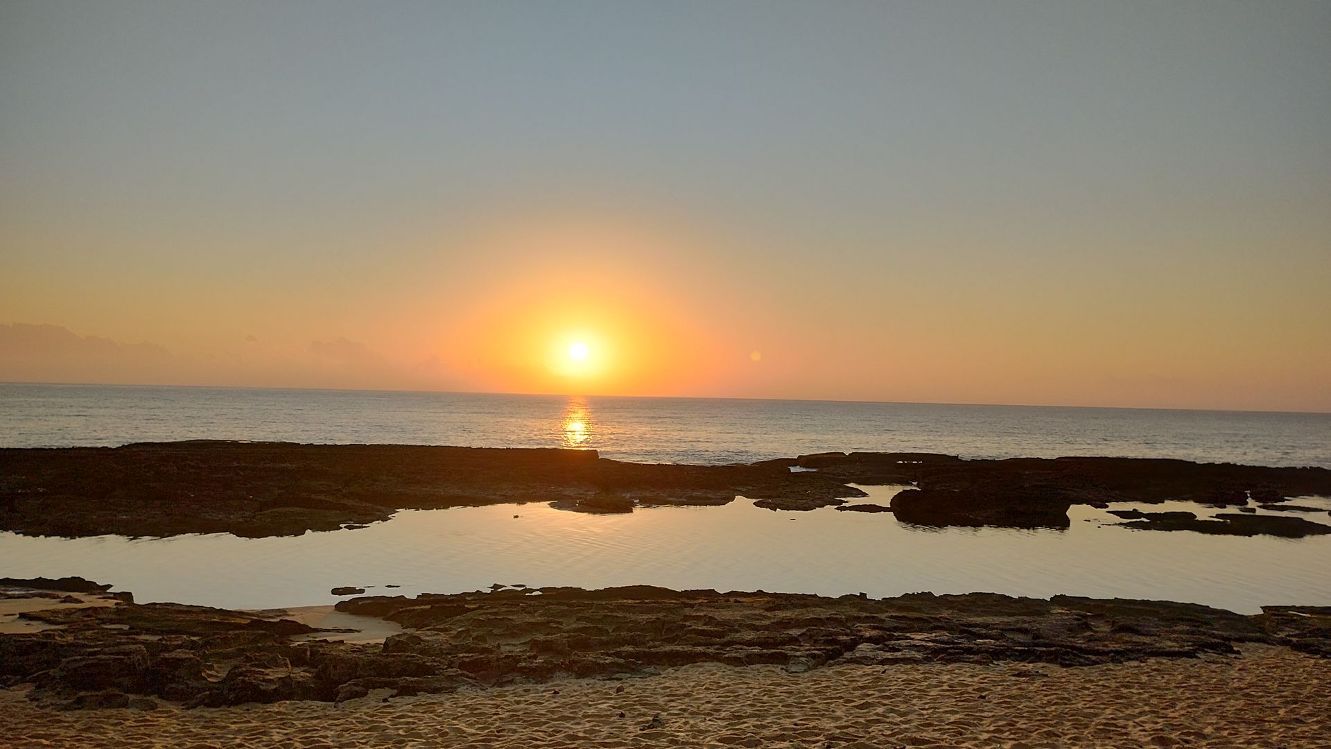 Pôr do sol sobre uma costa rochosa tranquila, com a luz dourada refletindo na água.