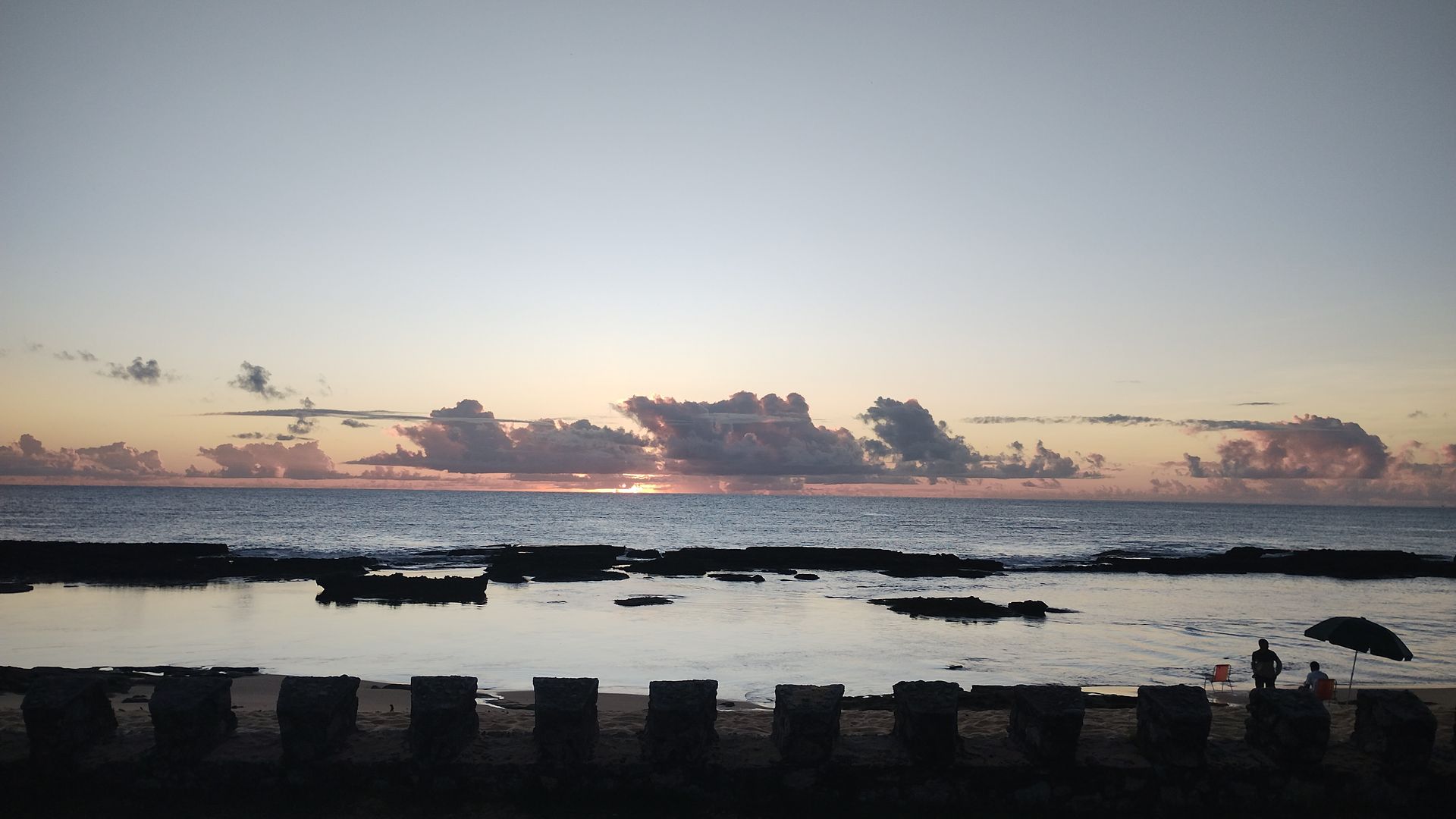 Pôr do sol sobre águas calmas com silhuetas de rochas e um céu nublado em tons de rosa e laranja.