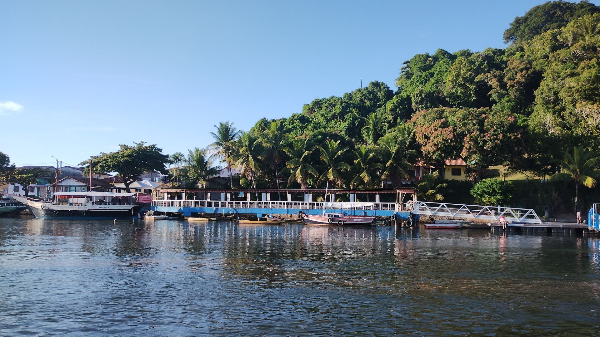 Barcos atracados ao longo de uma margem tranquila, ao lado de uma encosta arborizada, sob um céu azul límpido.