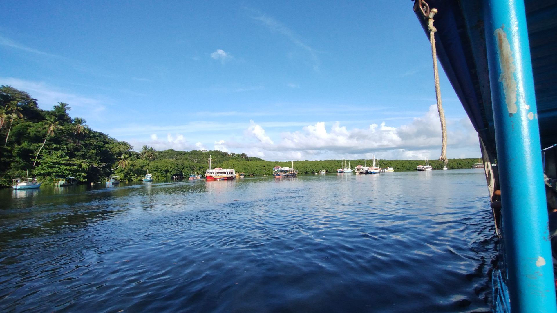 Rio calmo com céu azul, árvores na margem e a borda de um barco azul em primeiro plano.