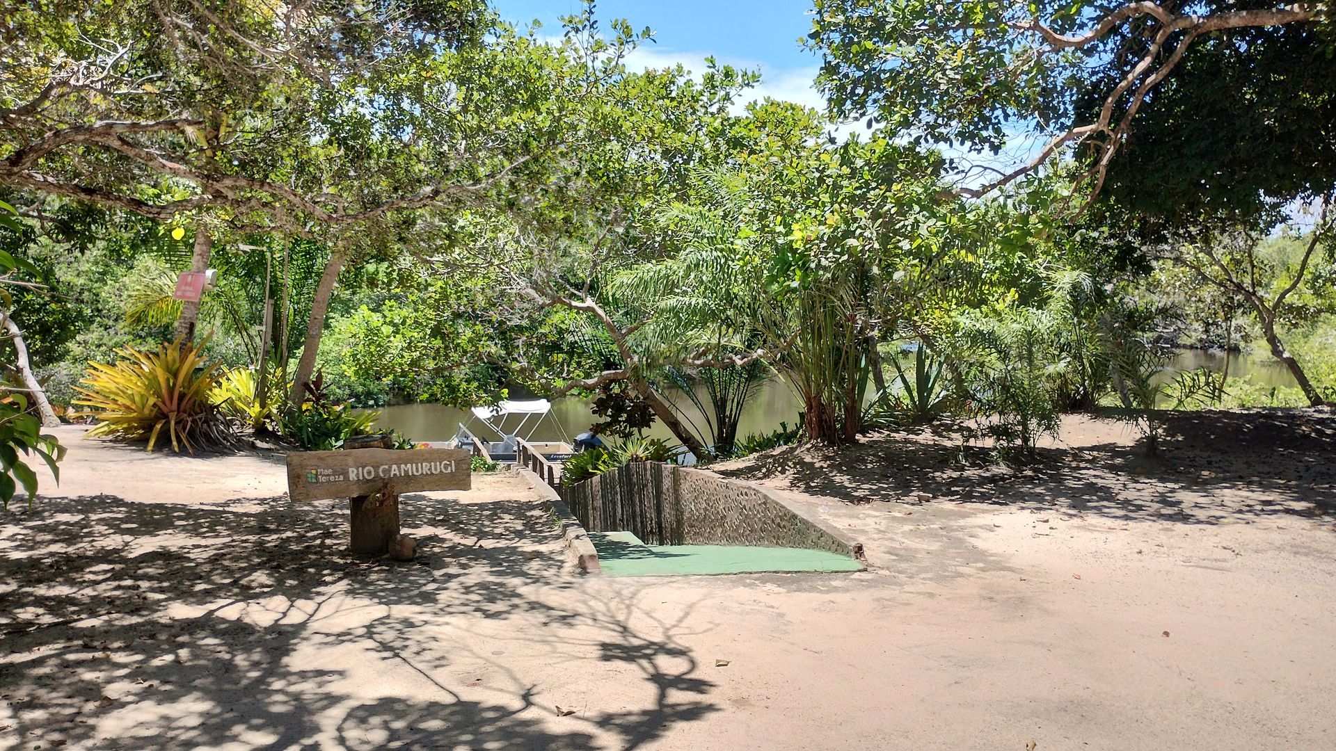 Caminho sombreado em um jardim externo com árvores, bancos de pedra e um tapete verde sob a luz do sol.