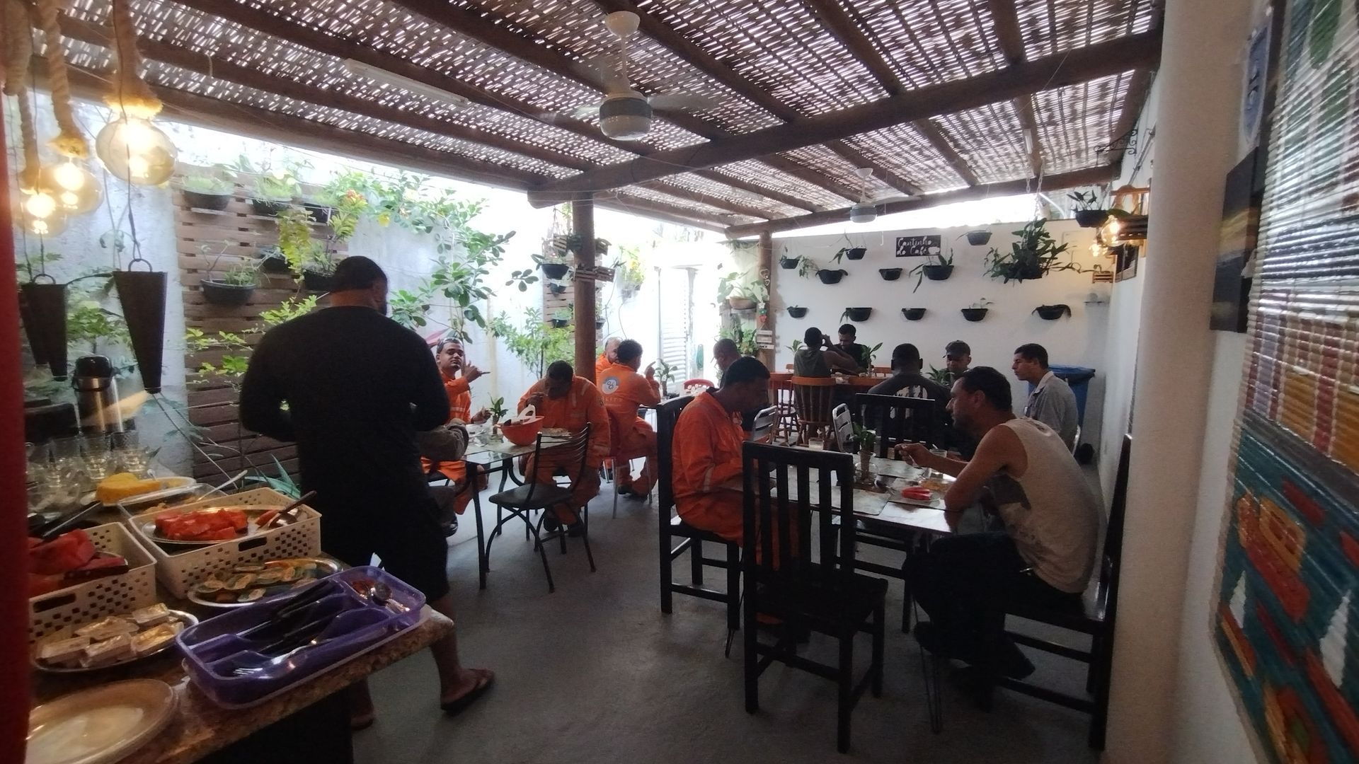 Área de refeições externa coberta, com mesas sob um teto de palha, ao lado de uma mesa de buffet.