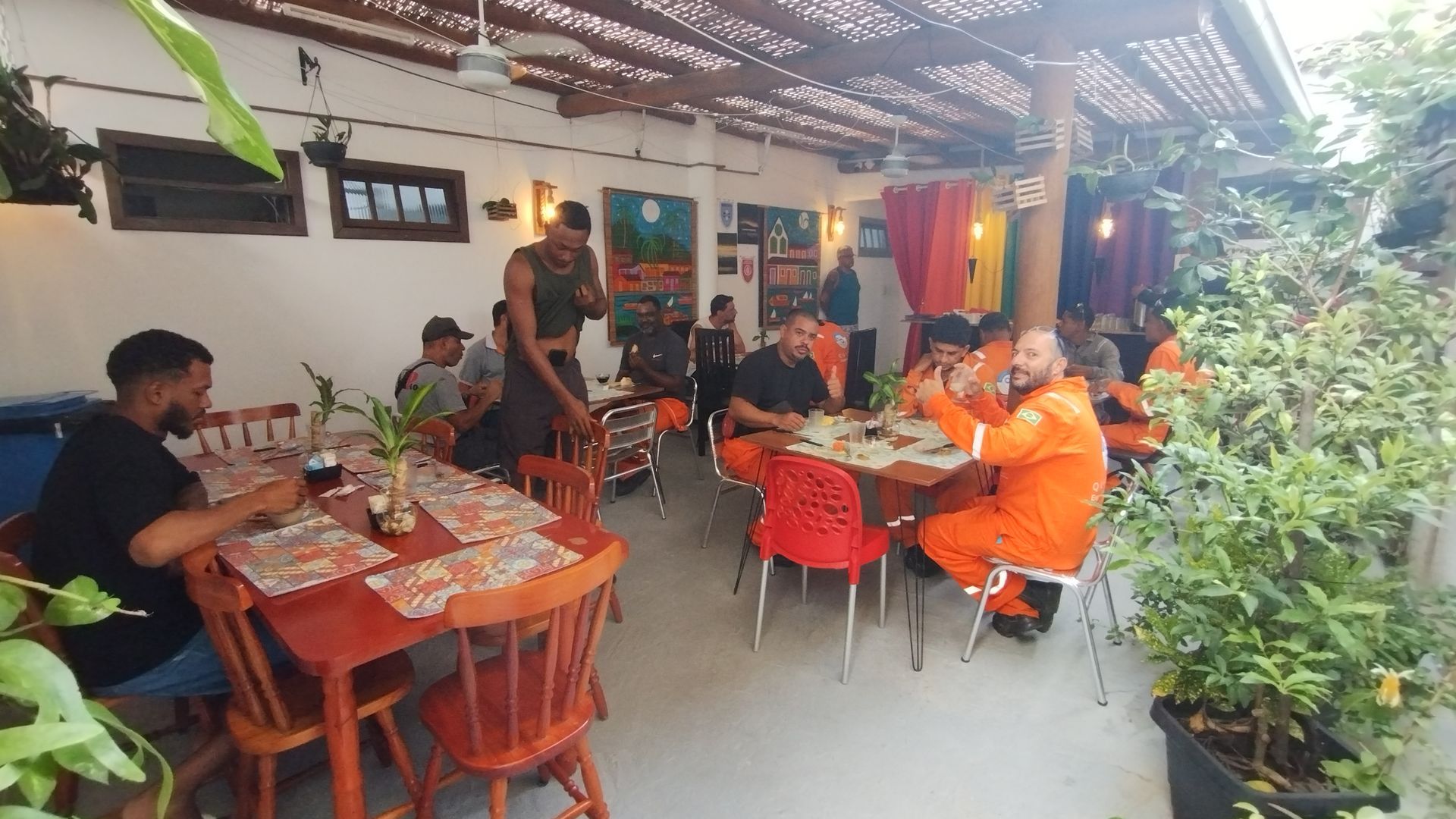 Restaurante ao ar livre com cadeiras laranjas, clientes jantando em mesas e vegetação exuberante do lado de fora.